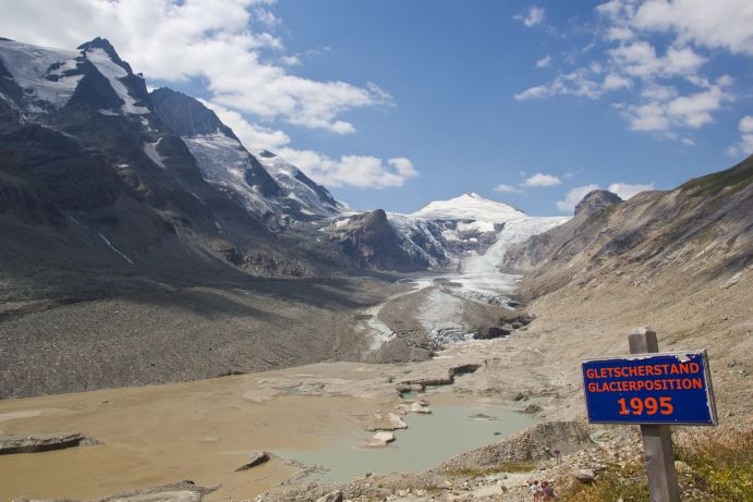 Im Vordergrund steht ein Schild mit der Aufschrift: &bdquo;Gletscherstand 1995&ldquo;. Im Hintergrund ist die weit zur&uuml;ckgegangene&nbsp;Pasterze zu sehen, der gr&ouml;&szlig;te Gletscher &Ouml;sterreichs am Fu&szlig;e des Gro&szlig;glockners.