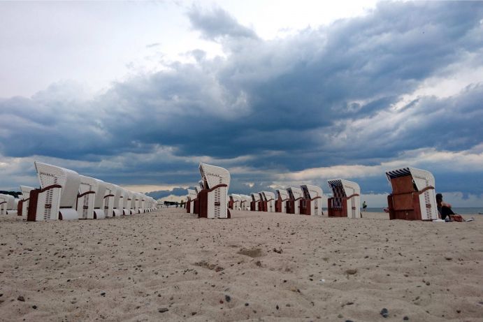 Strandk&ouml;rbe am Strand von Warnem&uuml;nde unter einem finsteren Wolkenhimmel. Eine Frau sitzt im Windschatten eines Strandkorbs und blickt auf die Ostsee hinaus.