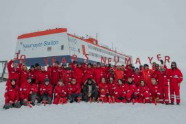 Gruppe von Menschen in roten Schneeanz&uuml;gen posiert vor Polarforschungsstation