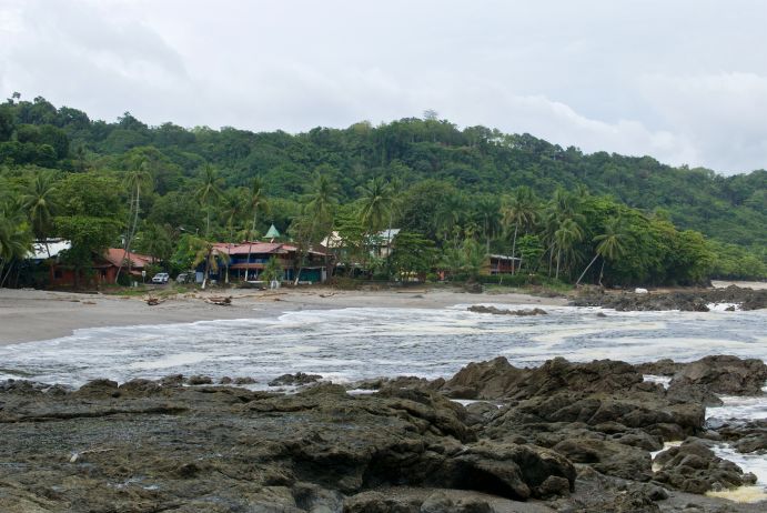 Ein tropischer Strand mit Felsen im Vordergrund, einem grünem Regenwald im Hintergrund, und Anlagen direkt am Rande des Strandes.