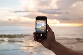 Person hält ein Smartphone mit Fokus auf das Meer und Wolken. 