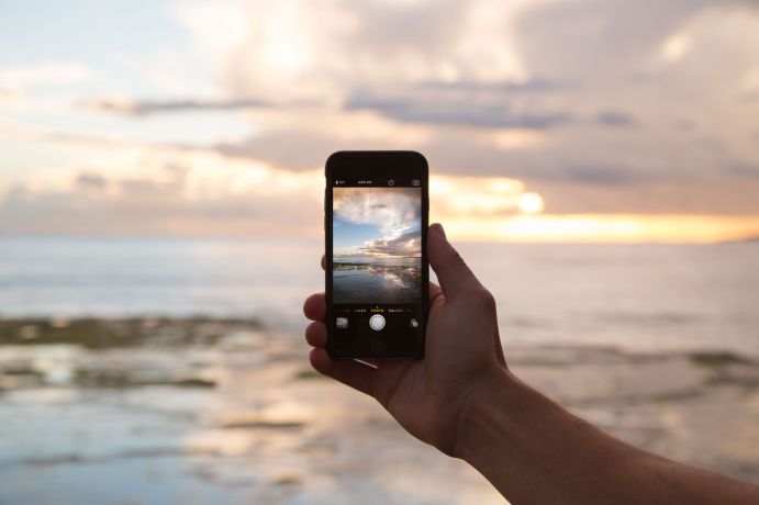 Person hält ein Smartphone mit Fokus auf das Meer und Wolken. 