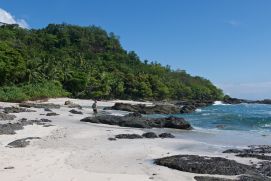 A person walks along a beach featuring some rocky outcrops. A tropical forest stretches along the background.