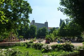 Das Bild zeigt den Klausenerplatz in Berlin-Charlottenburg. Man sieht einen Spielplatz in der Sonne mit Bäumen und einer Grünfläche mit blühenden Pflanzen im Vordergrund. 