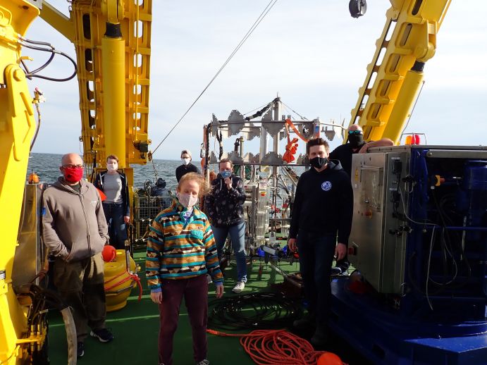 Expeditionsmitglieder mit Mund-Nasen-Schutz an Deck der Elisabeth Mann Borgese. Im Hintergrund ist der Lander BIGO1-01 des GEOMAR zu sehen, der Stoffflüsse an der Sediment-Wasser-Grenze misst. Forschungsfahrt während der Corona-Pandemie