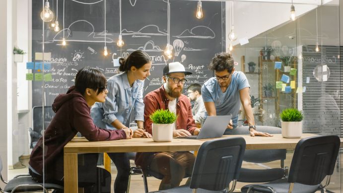 Junge Menschen gemeinsam an Tisch mit Laptop, im Hintergrund Tafel