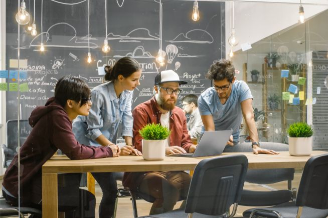 Junge Menschen gemeinsam an Tisch mit Laptop, im Hintergrund Tafel