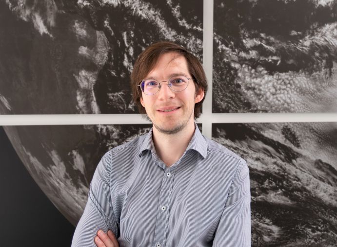 Dr. Mariano Mertens vor schwarz-weißem Satelittenbild der Erde. Er forscht am DLR-Institut für Physik der Atmosphäre in Oberpfaffenhofen und ist Nachwuchsgruppenleiter des Projektes IMPAC2T aus der Fördermaßnahme „BMBF-Nachwuchsgruppen Globaler Wandel: Klima, Umwelt und Gesundheit“.