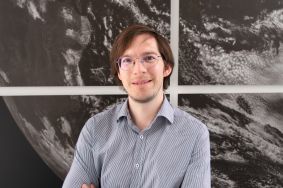 Dr. Mariano Mertens in front of a black-and-white satellite image of the Earth. He conducts research at the DLR Institute of Atmospheric Physics in Oberpfaffenhofen and is a junior research group leader of the IMPAC2T project from the funding measure "BMBF Junior Research Groups Global Change: Climate, Environment and Health".