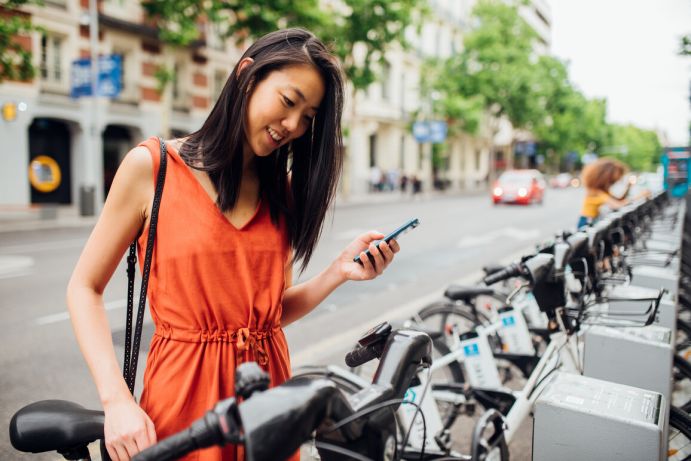 Junge fröhliche Frau, die mit dem Smartphone in der Hand das Fahrrad aus dem Fahrradträger auf der Straße nimmt