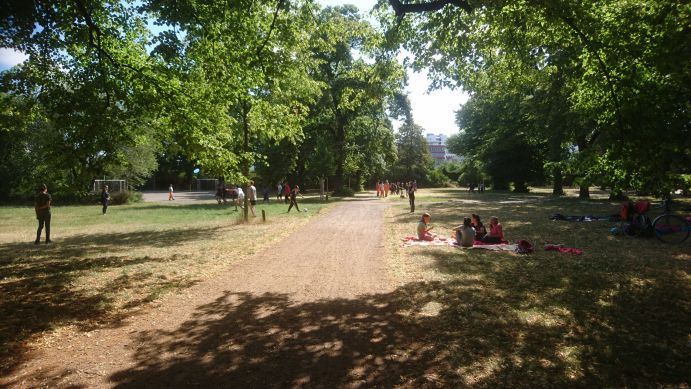 Friedenspark Leipzig im Sommer. Das Gras ist vertrocknet, Kinder sitzen auf einer Picknick-Decke oder spielen Fußball.